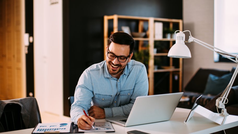 Young male professional in glasses and denim button down writing notes in a modern home office at a desk with a white top